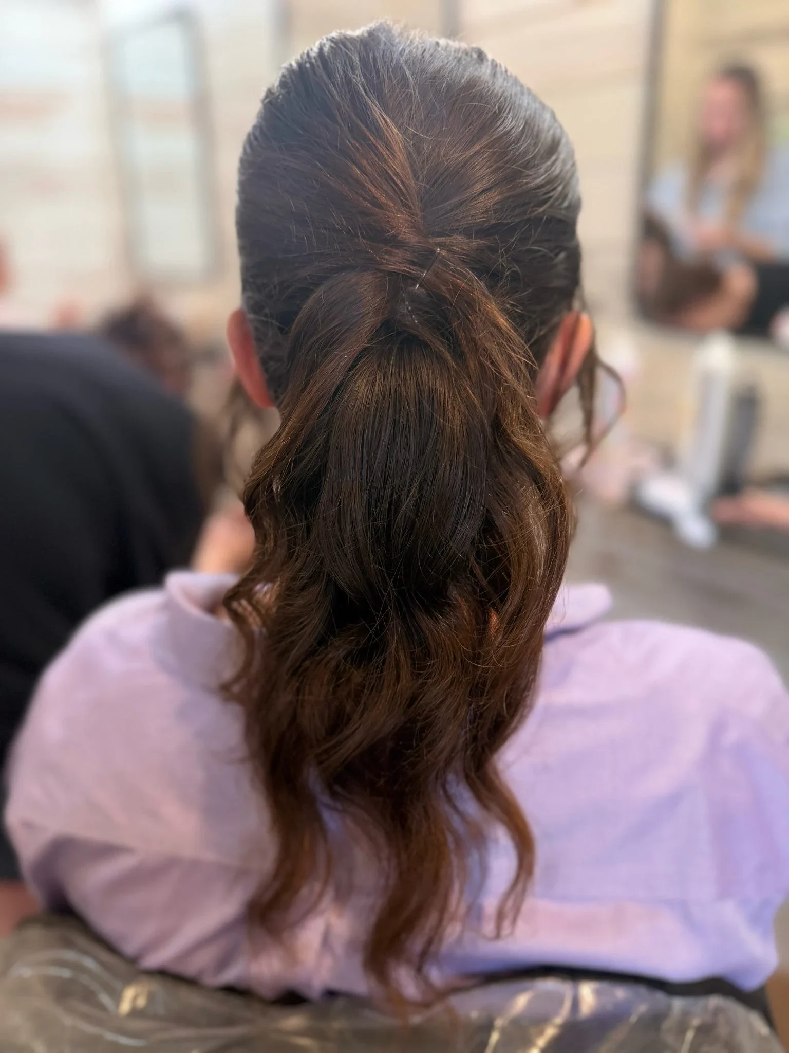 Back view of a brunette twisted low ponytail with curled ends styled by Glow Salon & Spa